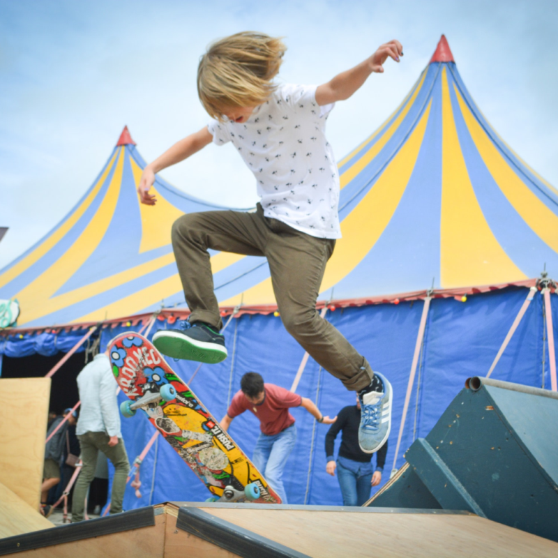 Ein Kind macht einen Trick auf einer Rampe auf dem Skateboard, im Hintergrund ist ein großes Zirkuszelt