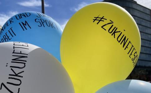 Mehrere Ballons sind aneinandergebunden. Mit schwarzem Stift sind Worte auf die Ballons geschrieben, die nicht alle zu erkennen sind. Lesbar ist das Wort "Zukünfte".