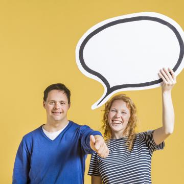 Eine junge Frau und ein junger Mann mit Down-Syndrom stehen fröhlich lachend nebeneinander. Sie hält eine Sprechblase über ihre Köpfe, er zeigt "Daumen hoch" mit seiner Hand.