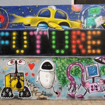 Das Bild zeigt ein Foto von einer Graffitiwand. In der Mitte des Graffitis steht groß Future. Im Hinergrund sind Hochhäuser, Planeten und ein fliegendes Auto. Im Vordergrund sind verschiedene Roboter zu sehen. 
