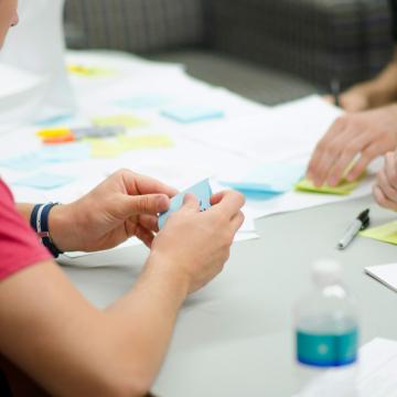 Personen sitzen an einem Tisch und halten ihre Ideen auf Papier und Post-its fest.