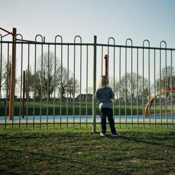 Kind steht vor Zaun, schaut auf Spielplatz.
