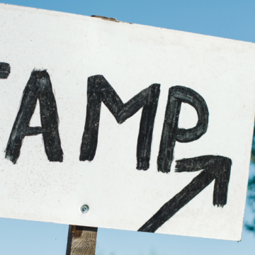 Auf einem handgemalten Schild steht "Camp" mit einem Pfeil nach vorne, der Himmel sieht sommerlich aus.