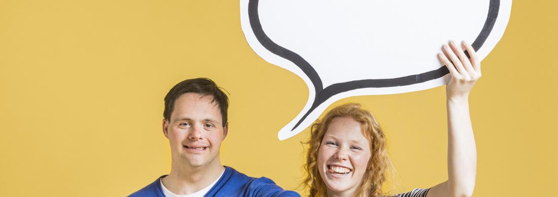 Eine junge Frau und ein junger Mann mit Down-Syndrom stehen fröhlich lachend nebeneinander. Sie hält eine Sprechblase über ihre Köpfe, er zeigt "Daumen hoch" mit seiner Hand.