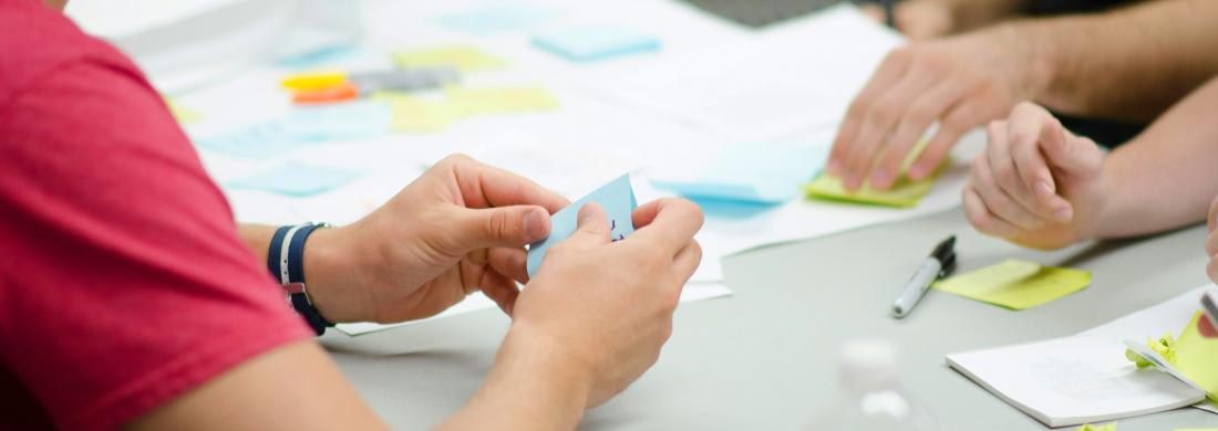 Personen sitzen an einem Tisch und halten ihre Ideen auf Papier und Post-its fest.