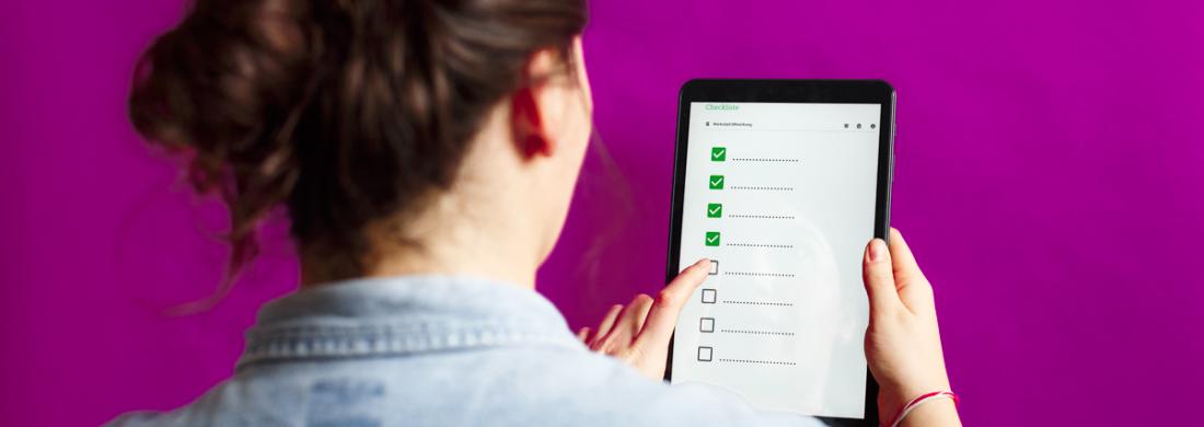 Blick von hinten über die Schulter einer Frau, die ein Tablet mit einer Checkliste in der Hand hält.
