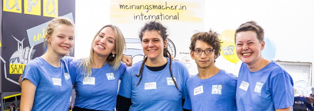 Fünf Jugendliche der meinungsmacher.innen stehen nebeneinander Arm in Arm und schauen in die Kamera. Alle haben ein hellblaues Meinungsmacher.in-T-Shirt an.