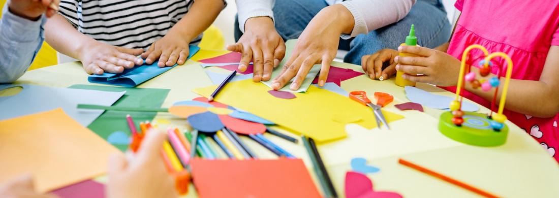 An einem Tisch basteln Kinder mit buntem Papier, Kleber, Schere und Stiften