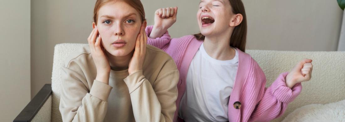 Eine junge Frau hält sich die Ohren zu, während ein junges Mädchen neben ihr laut ist und groß gestikuliert.