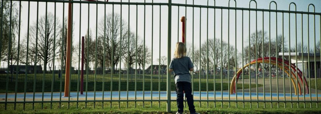 Kind steht vor Zaun, schaut auf Spielplatz.