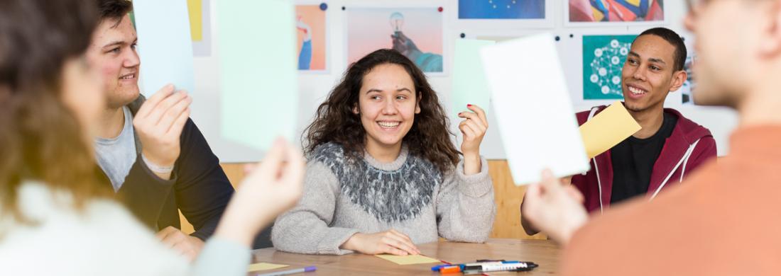 Junge Menschen sitzen an einem Tisch, halten Kärtchen hoch und lächeln