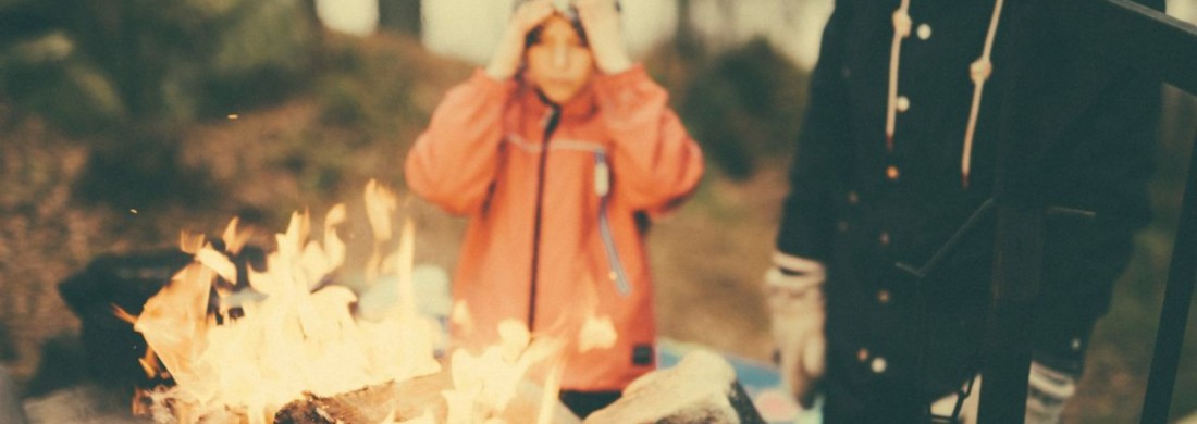 Zwei Kinder stehen draußen an einem Lagerfeuer; eines mit orangefarbener Jacke blickt auf die Flammen, das andere steht daneben.
