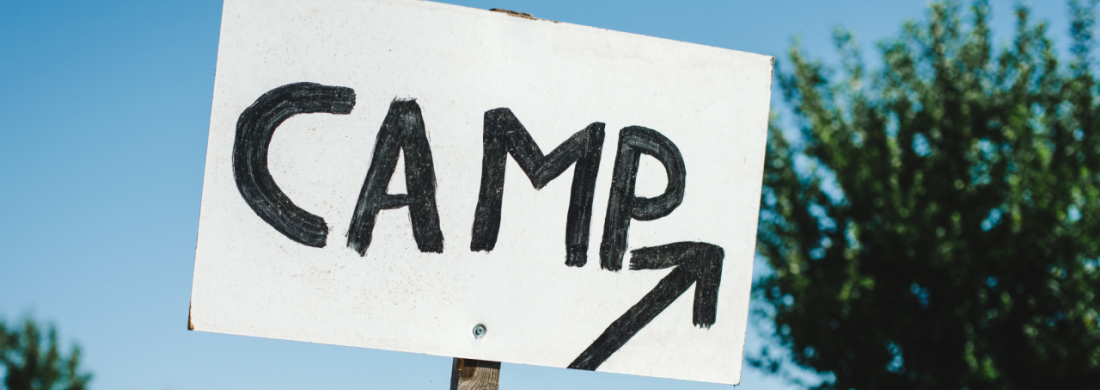 Auf einem handgemalten Schild steht "Camp" mit einem Pfeil nach vorne, der Himmel sieht sommerlich aus.