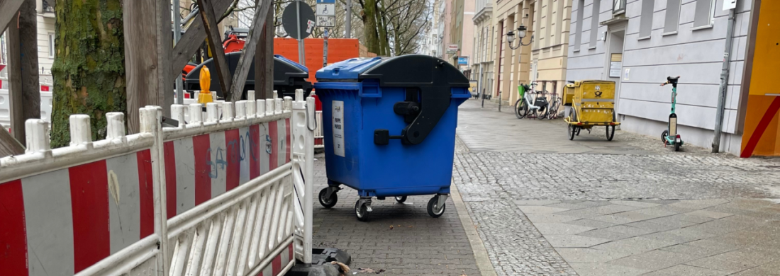 Müllcontainer blockiert schmalen Radweg, auf den ein Bauzaun ragt.