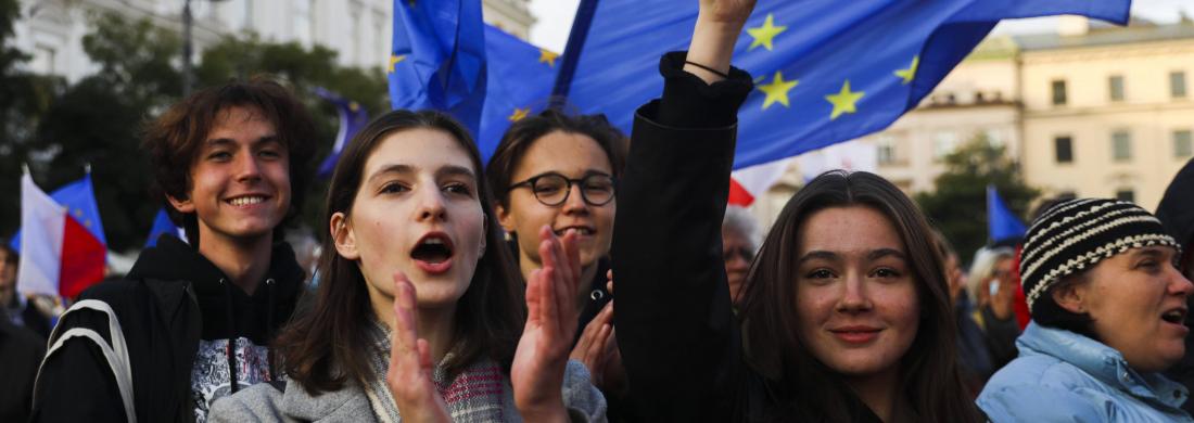 Im Fokus stehen vier Jugendliche bei einer Demonstration mit vielen Menschen im Hintergrund. Die Jugendlichen schwenken die europäische Fahne, eine Jugendliche ruft etwas und klatscht dabei in die Hände.