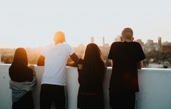Vier junge Menschen von hinten stehen auf einer Aussichtsplattform und blicken über eine Großstadt im Abendhimmel