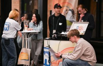 Mehrere Personen beim Politischen Abend des Landesjugendring Berlin: Ein junger Mann liest ein Dokument, während andere sich unterhalten oder an einem Tisch sitzen; im Hintergrund ein Banner der meinungsmacher:innen.