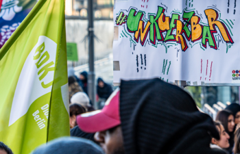 Bild einer Demo mit unterschiedlichen Fahnen und einem Plakat im Fokus, auf dem #unkürzbar steht