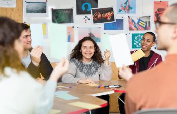 Junge Menschen sitzen an einem Tisch, halten Kärtchen hoch und lächeln