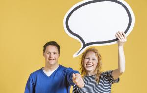 Eine junge Frau und ein junger Mann mit Down-Syndrom stehen fröhlich lachend nebeneinander. Sie hält eine Sprechblase über ihre Köpfe, er zeigt "Daumen hoch" mit seiner Hand.