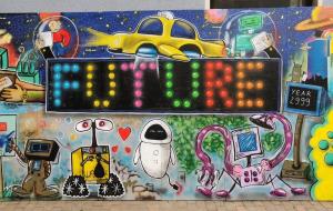 Das Bild zeigt ein Foto von einer Graffitiwand. In der Mitte des Graffitis steht groß Future. Im Hinergrund sind Hochhäuser, Planeten und ein fliegendes Auto. Im Vordergrund sind verschiedene Roboter zu sehen. 