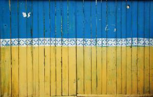 Ein Holzzaun, der in den Farben der ukraininschen Flagge gestrichen ist. Die obere Hälfte ist blau, die untere Hälfte gelb angestrichen