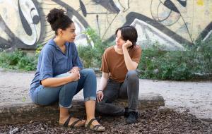 Eine Betreuerin und eine jugendliche Person unterhalten sich über etwas ernstes. Sie sitzen auf einem Baumstamm, der auf dem Boden liegt, im Hintergrund ist eine Wand mit Graffiti zu erkennen.