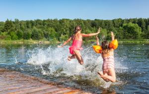 Zwei Mädchen springen von einem Holzplateau in einen See. Eine von ihnen trägt Schwimmflügel, im Hintergrund ist grüner Wald zu sehen, das Bild sieht sehr sommerlich aus.