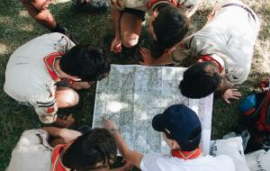 Sicht von oben drauf: Fünf Jugendliche beugen sich über eine Wanderkarte, eine Person zeigt auf einen Punkt auf der Karte.