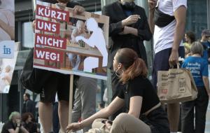 Eine Frau sitzt auf dem Boden und hält das Schild eines jungen Mannes, auf dem 'Kürzt uns nicht weg!' steht. Im Hintergrund sind weitere Demonstrationsteilnehmer zu sehen.