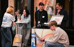 Mehrere Personen beim Politischen Abend des Landesjugendring Berlin: Ein junger Mann liest ein Dokument, während andere sich unterhalten oder an einem Tisch sitzen; im Hintergrund ein Banner der meinungsmacher:innen.