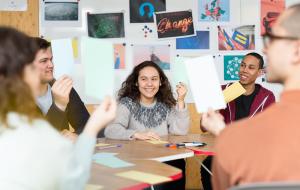Junge Menschen sitzen an einem Tisch, halten Kärtchen hoch und lächeln