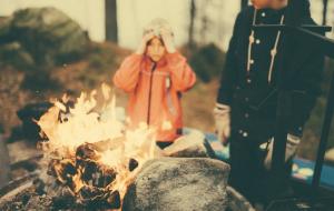 Zwei Kinder stehen draußen an einem Lagerfeuer; eines mit orangefarbener Jacke blickt auf die Flammen, das andere steht daneben.