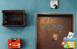 Tür mit Aufschrift "Büro", daneben ein Erste-Hilfe-Kasten, offene Stromleitungen und ein Schild auf dem "Because I'm worthless / Weil ich wertlos bin" steht)